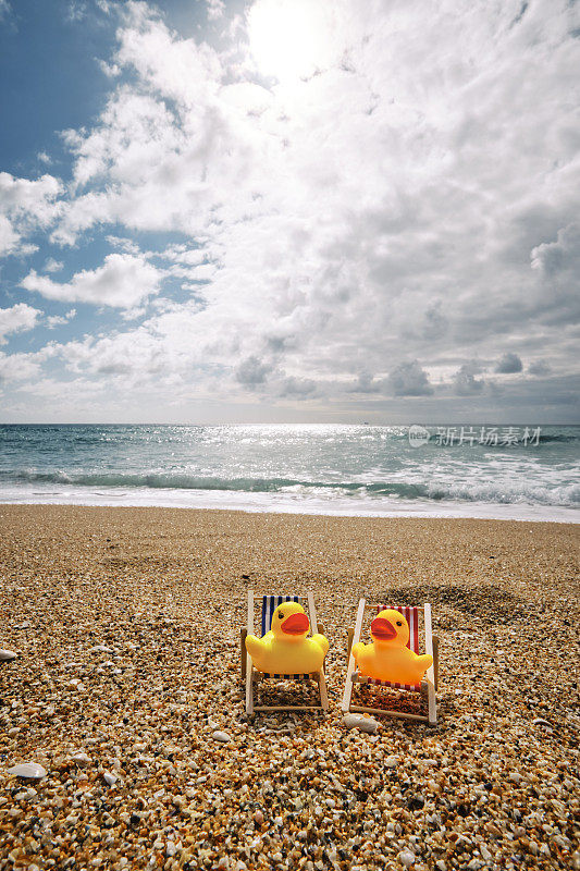 在一个晴朗的多云/阳光明媚的日子里，两只橡皮鸭坐在康沃尔波思科诺海滩的迷你躺椅上。